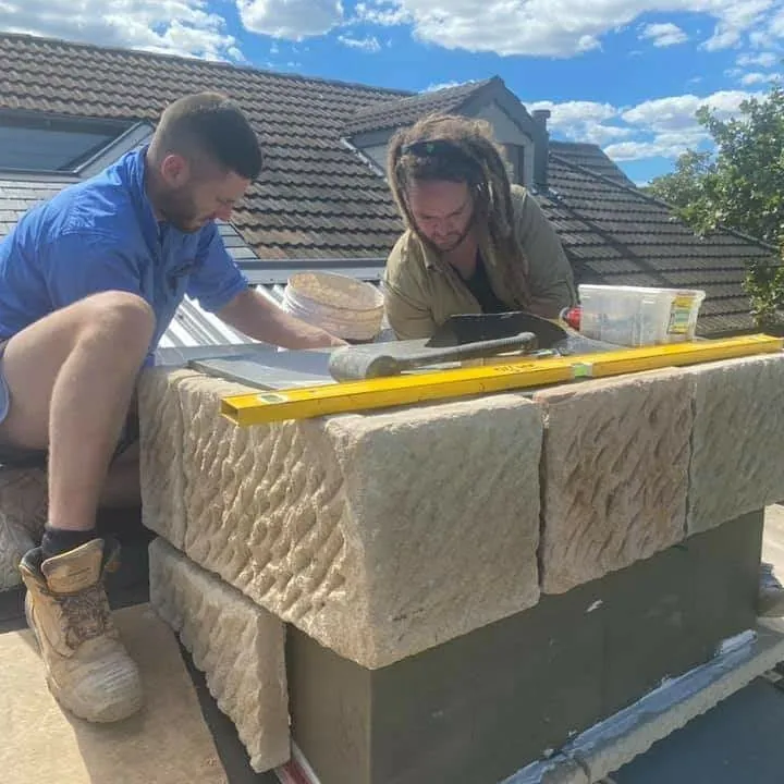 Two stonemasons building sandstone chimney on rooftop
